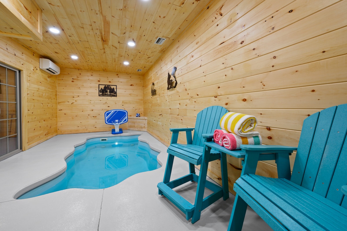 An indoor heated swimming pool is featured in a cozy log cabin room, surrounded by smooth concrete flooring and warmly hued wooden walls. Two aqua-blue chairs are positioned nearby, with neatly rolled towels in bright colors resting on one chair.