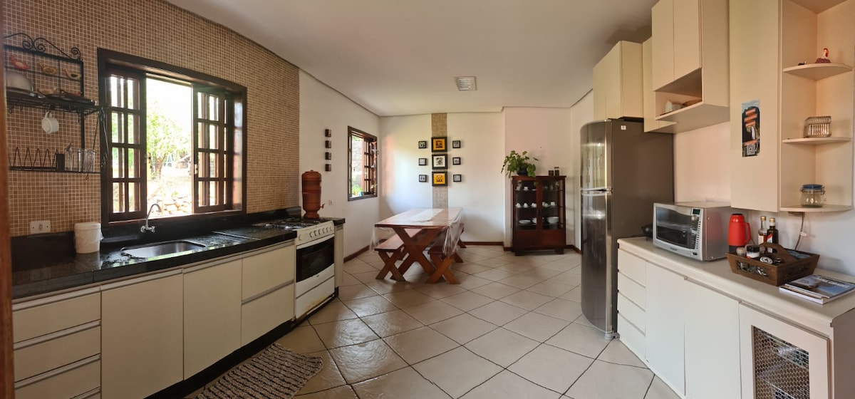 A spacious kitchen is featured, equipped with modern appliances and ample countertop space. A wooden dining table with cross-legged design is positioned near large windows, allowing natural light to fill the room. Shelving displays decorative items and potted plants, enhancing the functional layout.