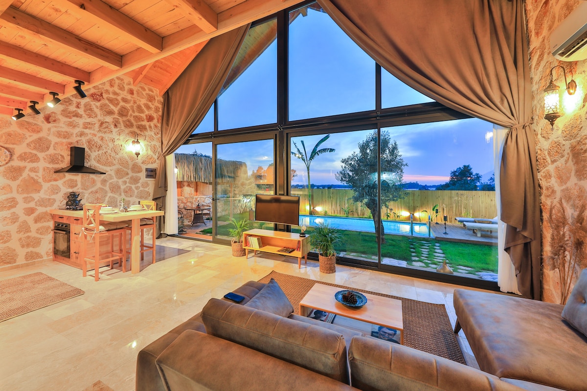 A spacious living area features large glass windows, allowing natural light to fill the room. A comfortable sectional sofa is complemented by a low coffee table. The view overlooks a well-maintained garden with a pool, framed by stone and wooden architecture.