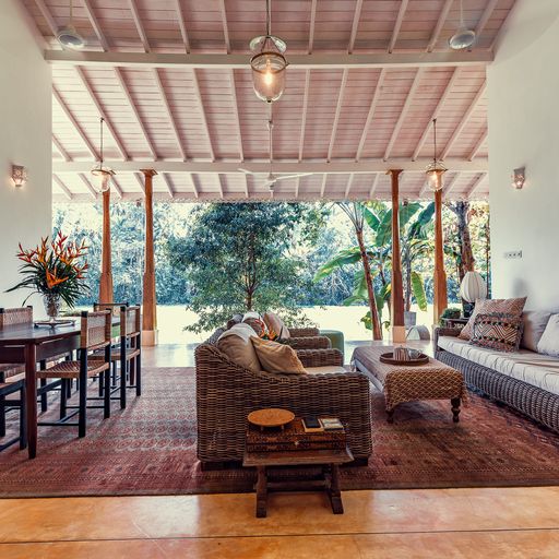 A bright living and dining area is seen, featuring woven furniture and a large sofa. Natural light filters through open spaces, illuminating the room. A dining table for six is positioned nearby, with views of the lush garden visible through the expansive windows.