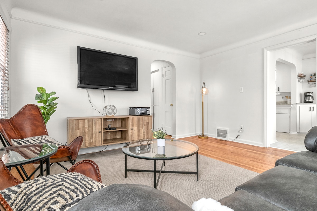 The living area features a comfortable seating arrangement with a stylish rug and a glass coffee table. A wall-mounted TV is positioned above a wooden media console. Natural light enters through the window, illuminating the space, while a decorative plant adds a touch of greenery.
