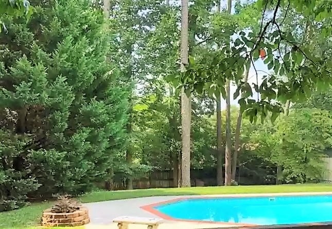 A vibrant backyard features a refreshing blue pool surrounded by lush greenery. Towering trees provide shade, while a brick fire pit is positioned nearby on the patio. The space offers a tranquil environment perfect for relaxation.