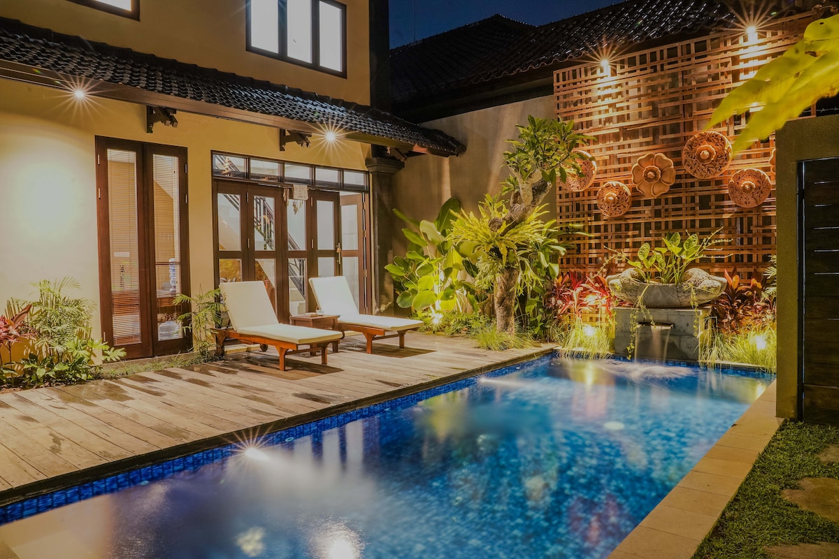 A serene pool area is presented, featuring two lounge chairs positioned beside a shimmering blue pool. Lush greenery surrounds the space, with decorative elements integrated into the walls, all softly illuminated by warm outdoor lighting.