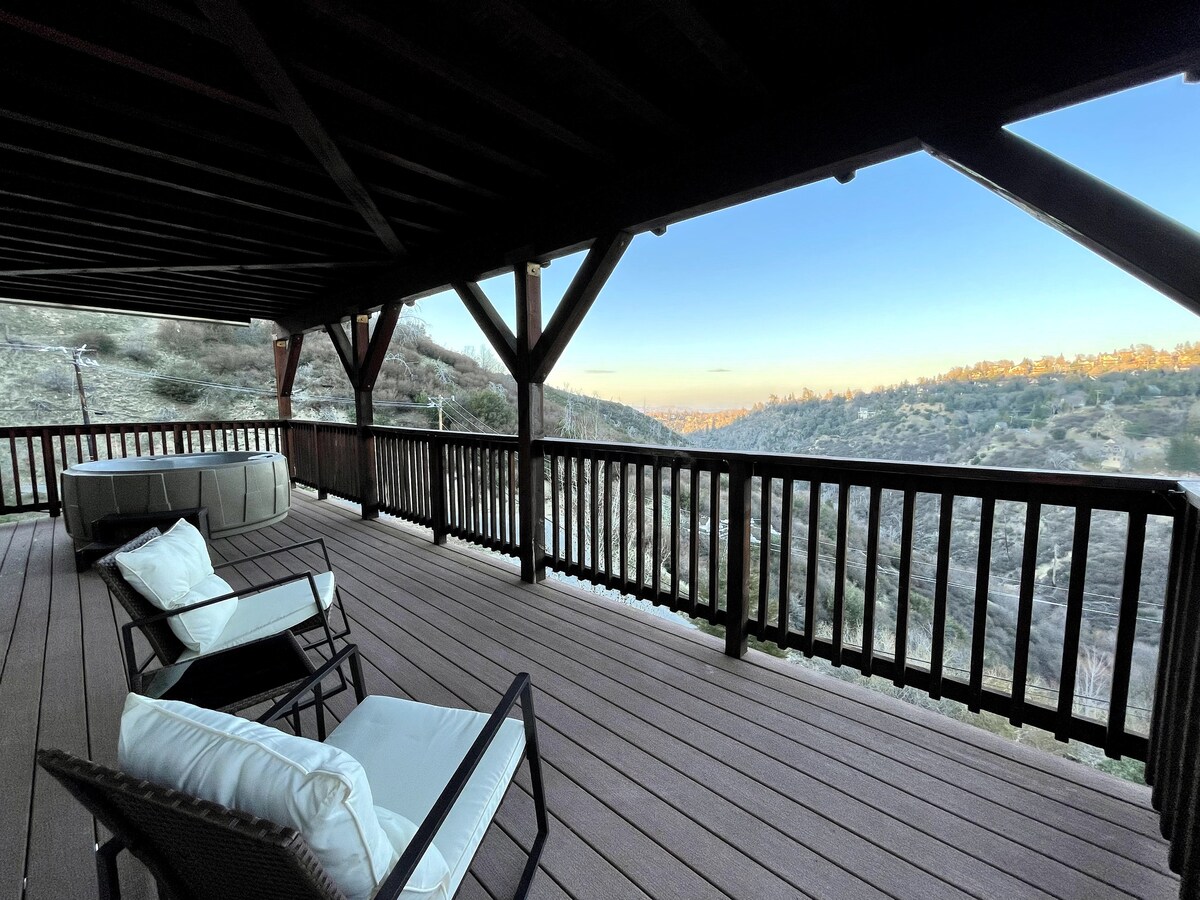 A covered deck is showcased with dark wooden beams and railings. Two lounge chairs with white cushions are positioned for relaxation, while a spacious seating area is visible. The scenic view of the landscape can be observed in the distance, enhancing the outdoor experience.