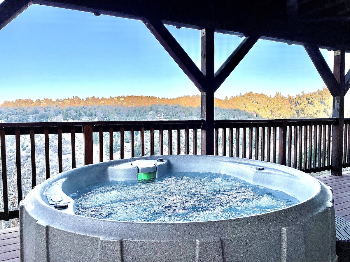 A relaxing hot tub is visible on a spacious wooden deck, framed by a rustic, covered structure. Wood railing offers a view of the surrounding landscape, highlighting the green hills in the distance under a clear sky.