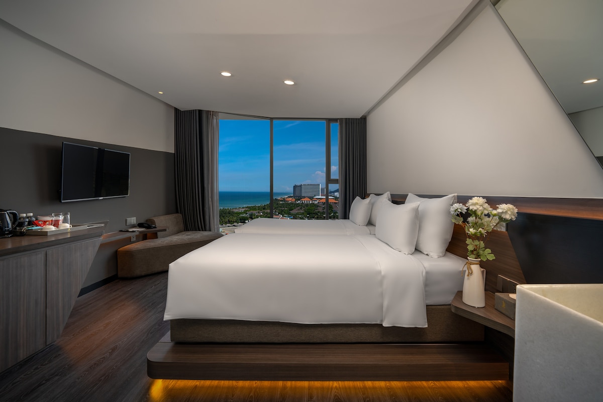 A modern guest room features two spacious beds with crisp white linens. A large window offers a view of the ocean and city landscape. A cozy seating area is situated near the entrance, and a television is mounted on the wall opposite the beds.