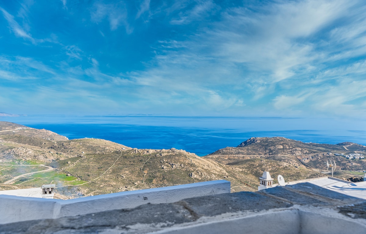 A sweeping view of the Aegean Sea stretches across the horizon, with rolling hills and rugged terrain visible in the foreground. The sky features varying shades of blue, creating a serene backdrop that enhances the natural beauty of the landscape.