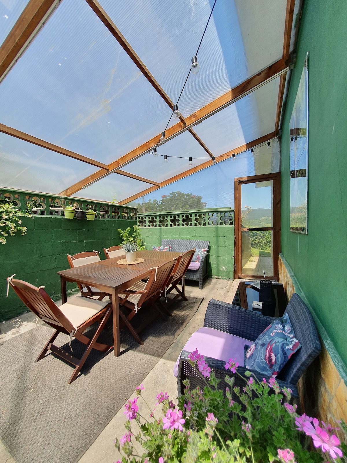 A covered outdoor seating area features a wooden dining table surrounded by six chairs, with colorful flowers enhancing the space. A lounge area is provided with black wicker seating. The green walls and ceiling create a lively atmosphere, while string lights add subtle illumination.