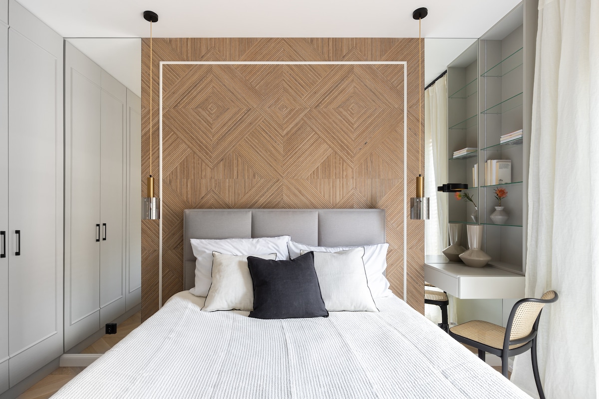 A modern bedroom is showcased with a textured wood wall accent behind a spacious bed. White linens and fluffy pillows are neatly arranged on the bed. Elegant pendant lights hang above. A light-filled nook features decorative shelves and a minimalistic chair.