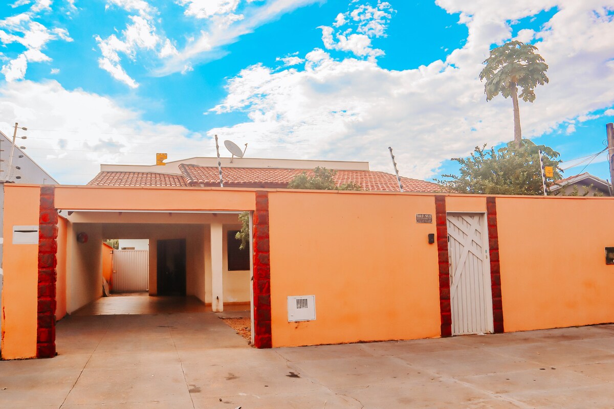 The exterior of the house is depicted, showcasing a bright orange façade with red stone accents. A spacious driveway is visible, leading to a gated entrance. The sky is partly cloudy, adding a vibrant backdrop to the scene.
