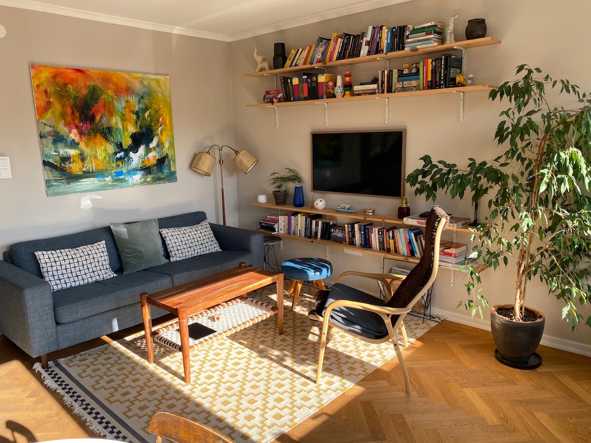 A living area is showcased with a navy blue sofa adorned with patterned cushions. A wooden coffee table is positioned in front, complemented by an accent chair. A bookshelf filled with various books is mounted above, and a flat-screen TV is visible on the wall. Natural light illuminates the room.