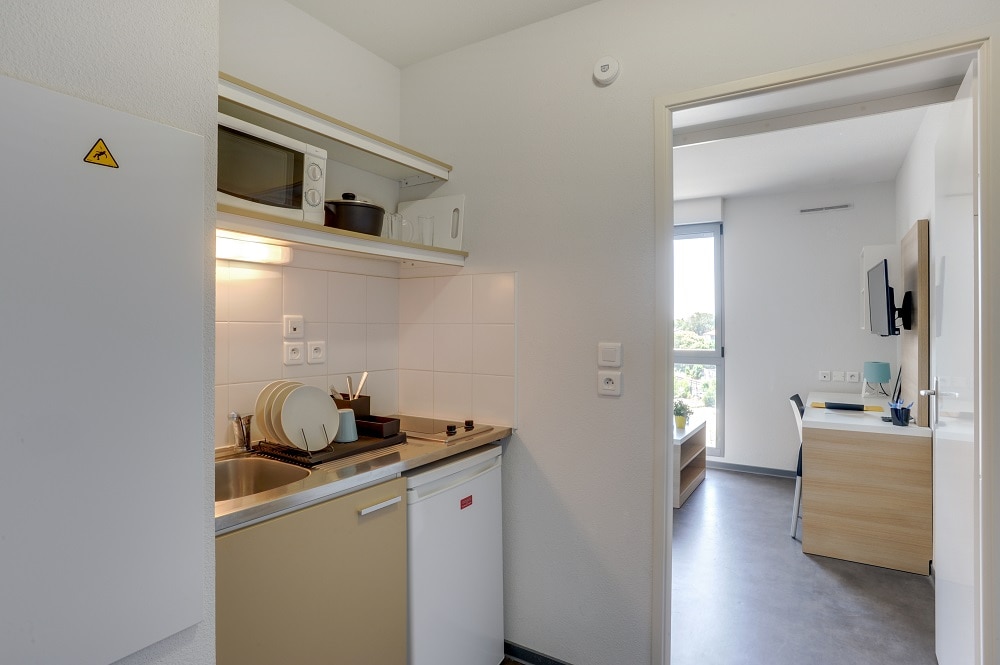A compact kitchenette is equipped with a sink, microwave, and refrigerator, providing essential cooking amenities. The doorway leads to a bright living area with natural light from a window, and a desk is visible in the adjacent room, ideal for working.