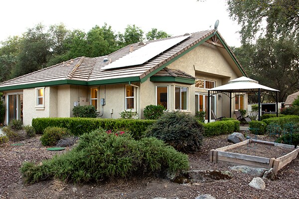 The exterior of the guest house is showcased, featuring a sloped roof with solar panels and large windows. Surrounding the structure, well-maintained shrubs and decorative rocks provide a natural aesthetic. A gazebo is visible in the backyard area, offering additional outdoor seating.