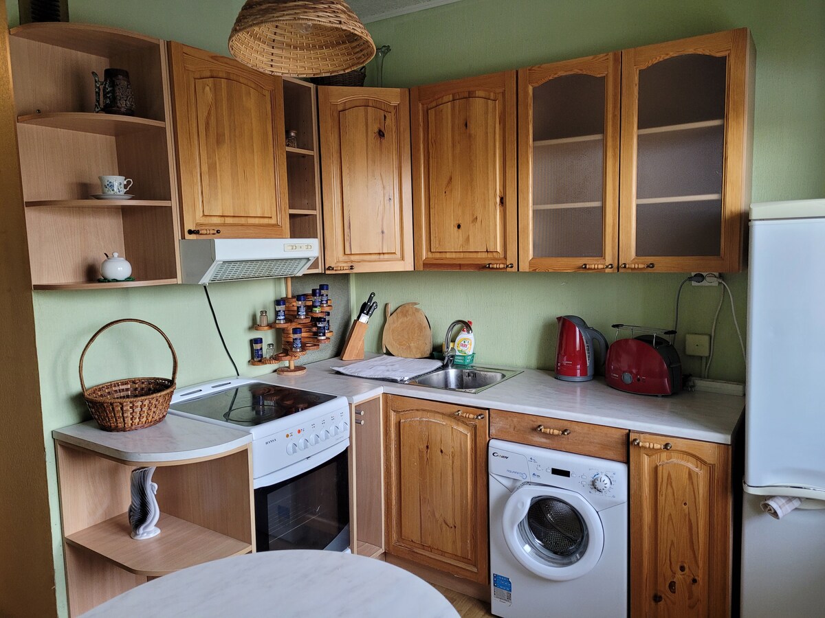 A functional kitchen is equipped with wooden cabinetry and modern appliances. The space includes an oven, stove, and refrigerator, along with a washing machine. Countertops are adorned with cooking utensils, and a woven basket provides additional storage.