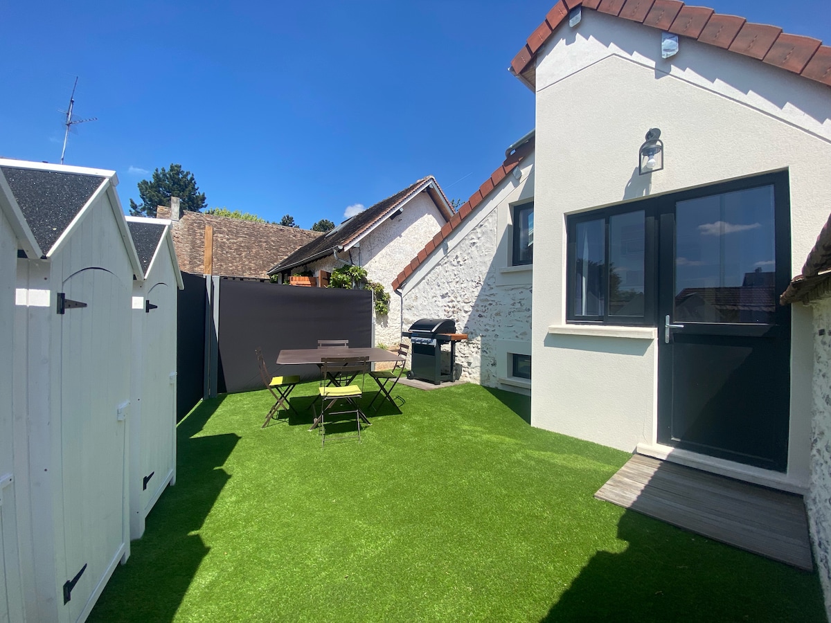 An outdoor area features a well-maintained green lawn, complemented by a table and chairs for dining. A barbecue grill is positioned nearby, and storage sheds are visible along the fence, providing a practical and inviting environment.