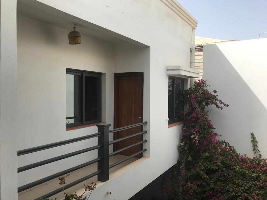 An exterior view of the apartment showcases a light-colored building with a wooden door and two windows. A railing borders a small balcony, while vibrant bougainvillea vines add color along the side, enhancing the serene environment.