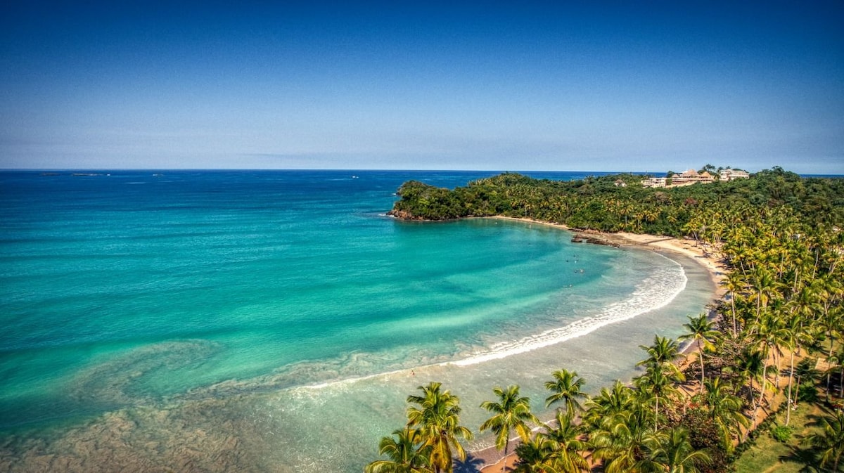 A scenic view of Playa Bonita showcases a beautiful curve of golden sand meeting turquoise waters, framed by lush palm trees. The tranquil beach is complemented by the gentle waves and a distant green hillside, creating a serene coastal atmosphere.