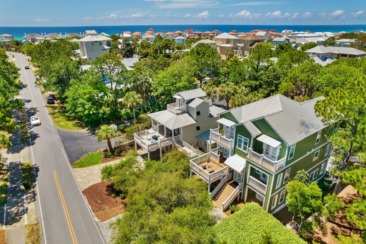 An aerial view captures the luxurious beach house surrounded by mature pine trees and greenery. The nearby coastline is visible in the background, with the ocean glimmering under the sunlight. The spacious deck and balconies invite outdoor relaxation, while the peaceful neighborhood streets complement the setting.