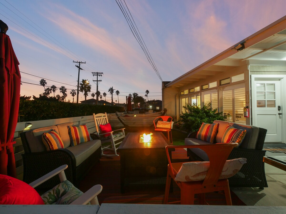 An outdoor patio is furnished with comfortable seating arrangements, including a large coffee table and a fire pit. Decorative cushions add color to the space, while palm trees can be seen in the background against a vibrant sunset sky.