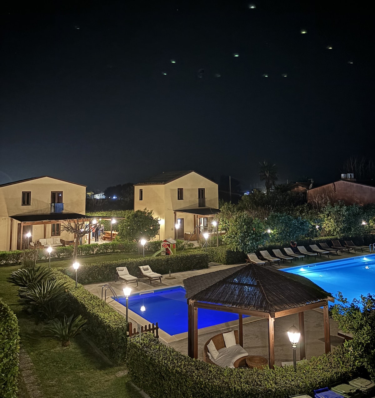 The nighttime view reveals a serene outdoor area featuring two illuminated swimming pools surrounded by lush greenery. Comfortable loungers are positioned near the pools. A thatched-roof gazebo is visible, providing a shaded space, while decorative lights enhance the tranquil atmosphere.