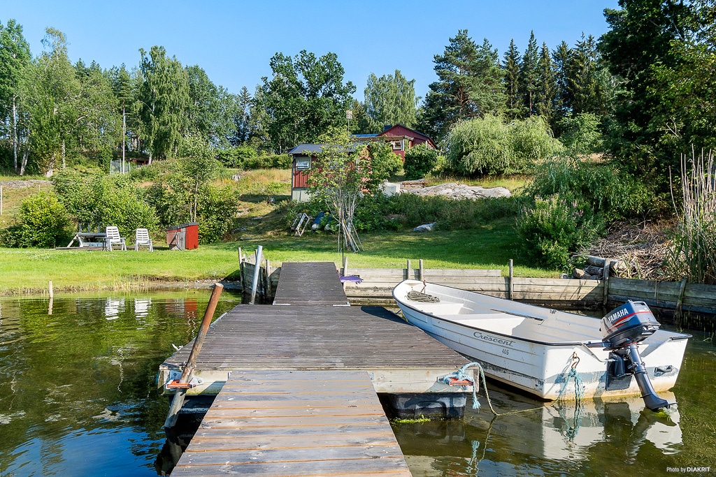 A wooden dock extends into calm waters, accommodating a boat moored alongside. Lush greenery surrounds the area, including trees and shrubs in the background. A red guesthouse and seating area can be seen on elevated land, framed by the natural landscape.