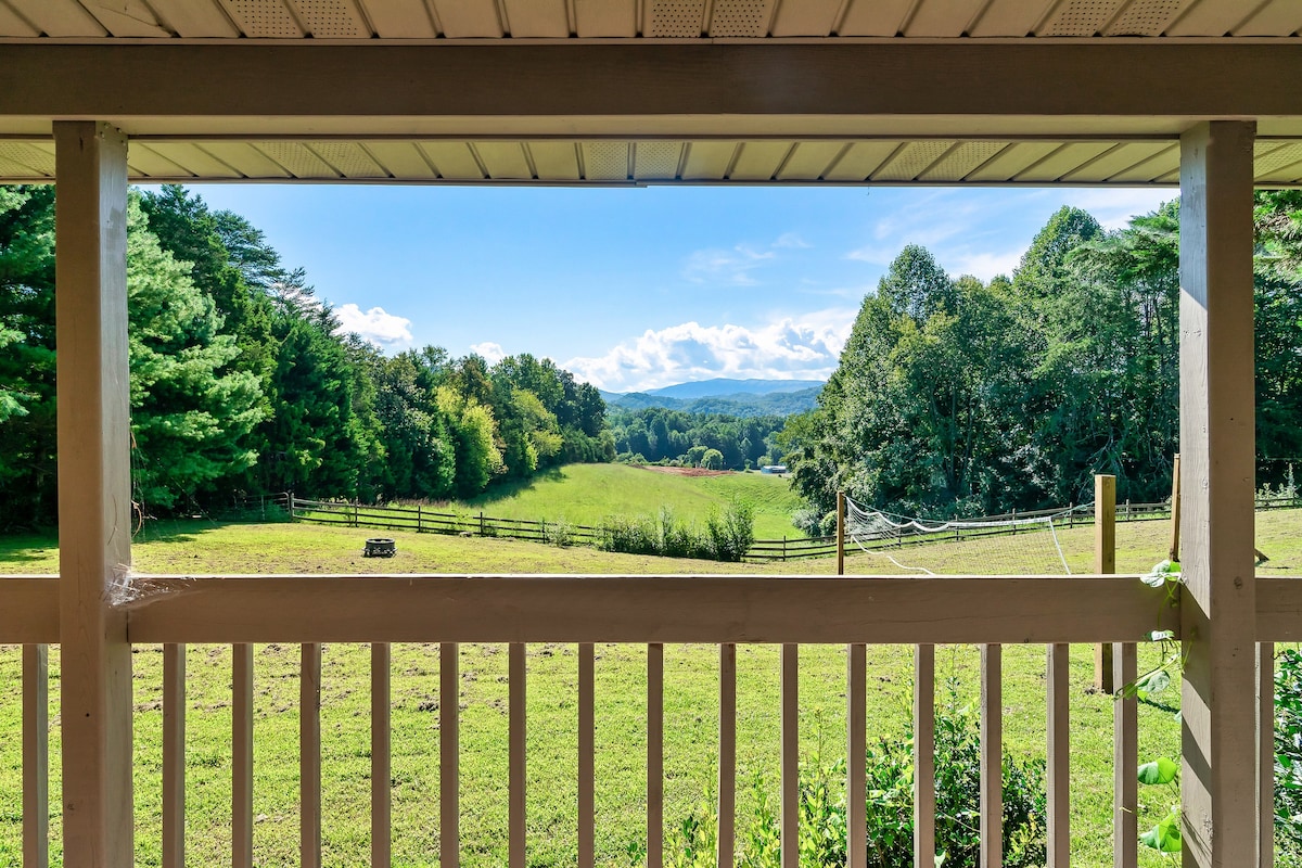A scenic view of rolling green hills and trees is framed by a wooden deck railing. Mountaintops can be seen in the background under a clear blue sky, creating a peaceful outdoor setting.