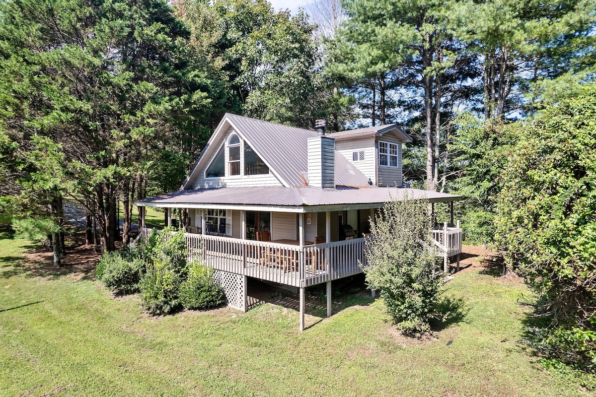 A cabin is nestled among trees on a spacious property, featuring a deck that wraps around the front. The sloped roof and multiple angles of the structure create an inviting profile. Lush greenery surrounds the exterior, enhancing the natural setting.