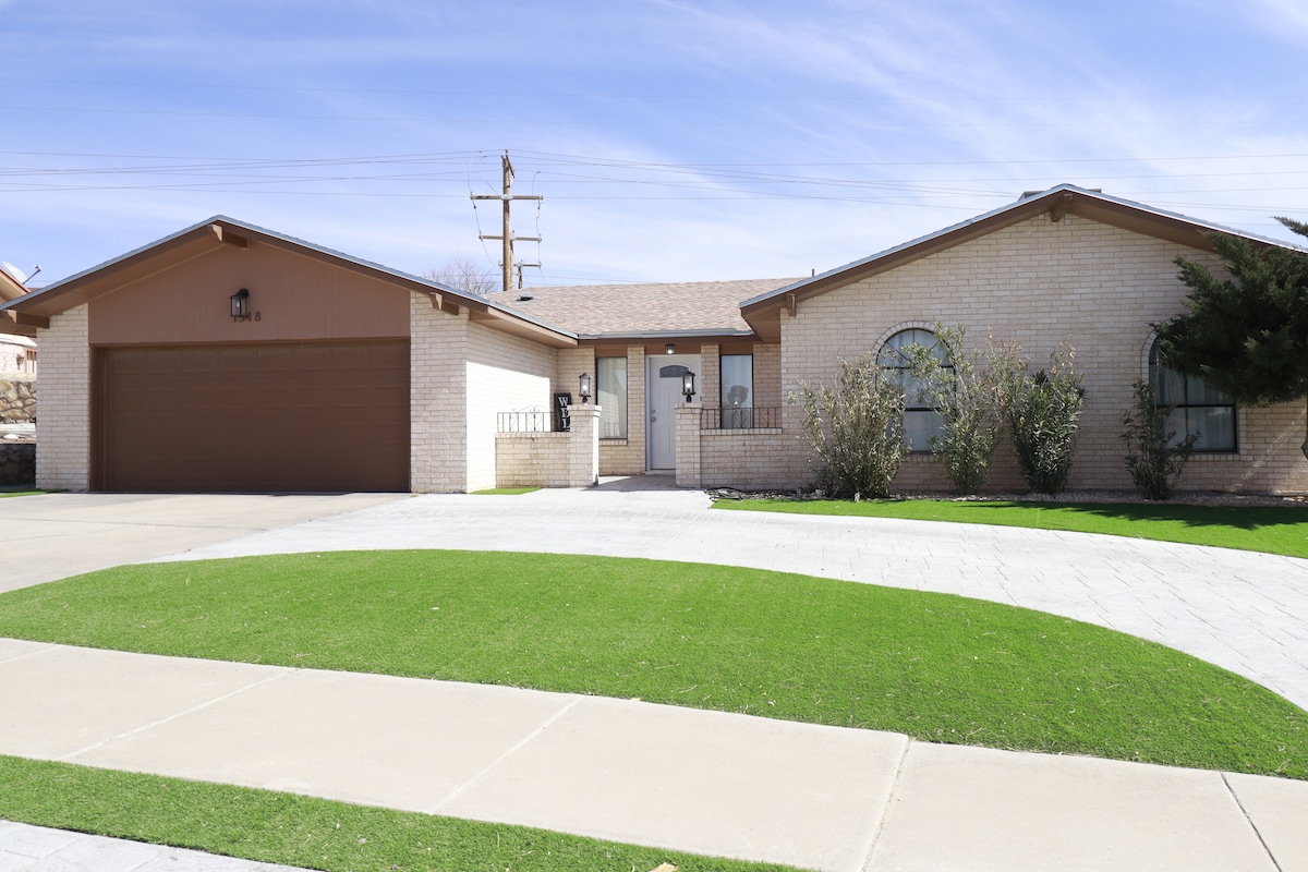 A single-story home with a brick facade is showcased, featuring a two-car garage and a landscaped front yard. A spacious driveway leads to a covered entrance, and large windows invite natural light into the residence. Green grass and trimmed shrubbery enhance the exterior appeal.