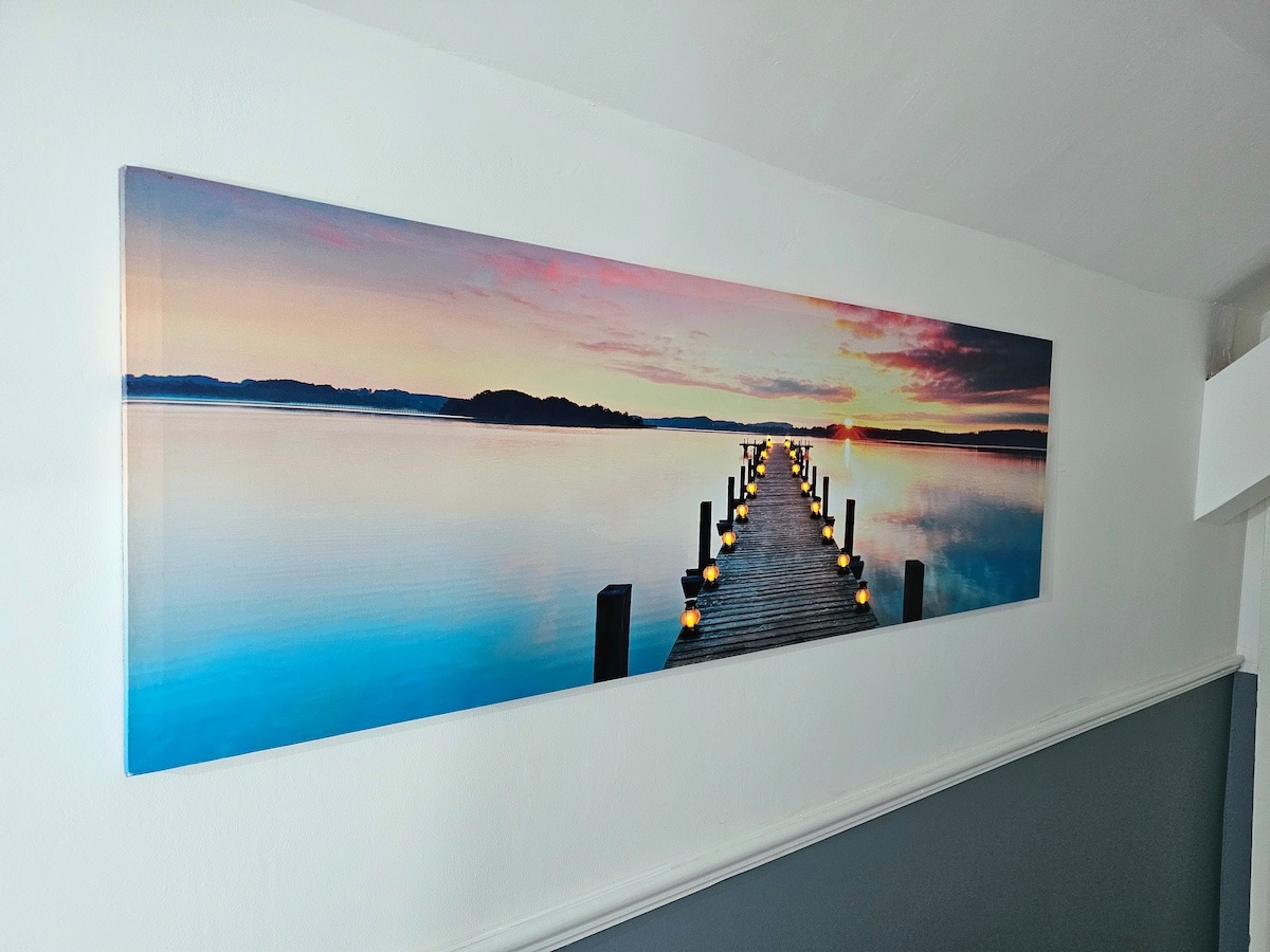 A long wall-mounted artwork depicts a serene waterscape at sunset, featuring a wooden pier extending into calm waters. Soft hues of pink and blue are visible in the sky, and lanterns line the edges of the pier, illuminating the scene with a gentle glow.