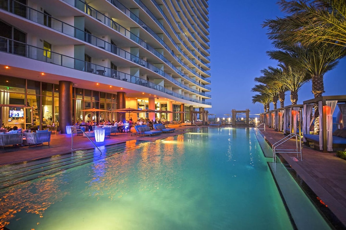 An expansive pool area is framed by modern lounge chairs and palm trees, illuminated by soft lighting that enhances the evening ambiance. The hotel’s structure looms in the background, with a vibrant atmosphere showcasing guests enjoying the space.