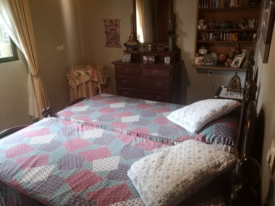 A cozy bedroom is presented with a wooden bed featuring a patchwork quilt. Two decorative pillows rest atop the spacious bed. A wooden dresser stands against the wall, complemented by a mirror and various decorative items on display. Natural light enters through a window with curtains.