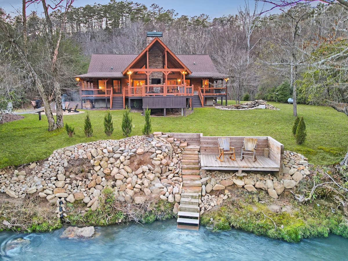 A log cabin, surrounded by trees, overlooks a gentle river. A spacious deck features multiple seating areas and a set of wooden chairs at the water's edge, with a staircase leading down to the riverbank. The grounds are landscaped with rocks and greenery.