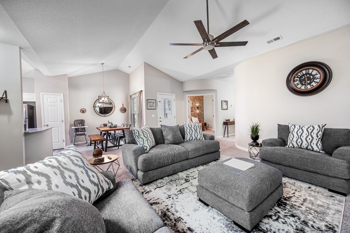 The living area features modern gray sofas arranged around a large central ottoman, with a light-patterned area rug underneath. High ceilings introduce a spacious feel, complemented by a ceiling fan. A dining area is visible in the background, showcasing a round table and high chair.