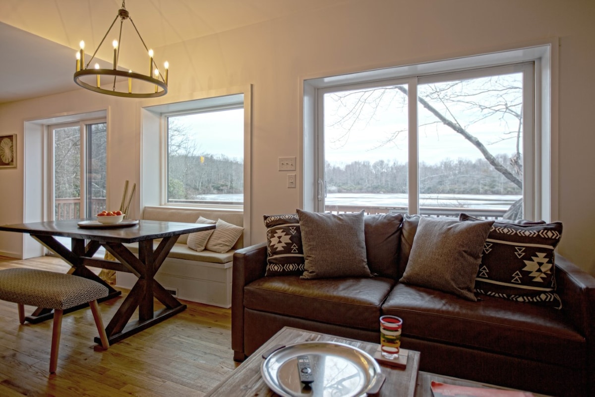 A cozy living area is highlighted by a brown sofa adorned with patterned throw pillows. A wooden dining table is set nearby, accompanied by a sleek chair. Large windows provide natural light and showcase views of the serene waterfront outside.