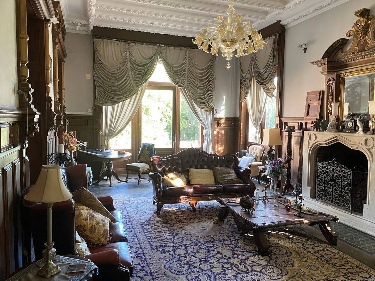 The living area features elegant drapery framing large windows, allowing natural light to fill the space. Antique furnishings, including a leather seating arrangement and a low wooden table, are complemented by a decorative fireplace. A rich area rug adds comfort to the hardwood floors.