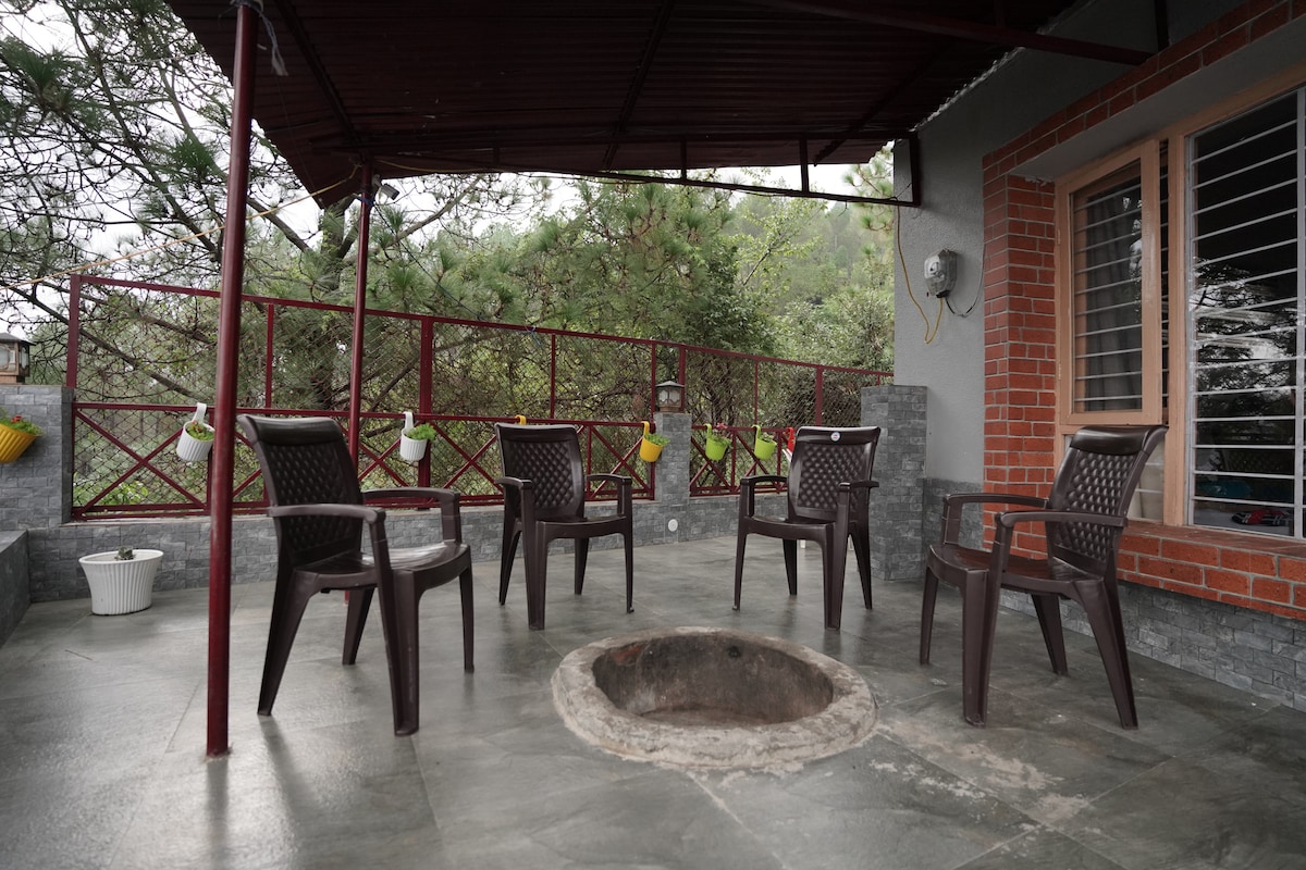 An outdoor seating area features six brown plastic chairs arranged around a circular stone fire pit. The space is shaded by a maroon roof and is surrounded by greenery, with a decorative fence providing a natural backdrop.