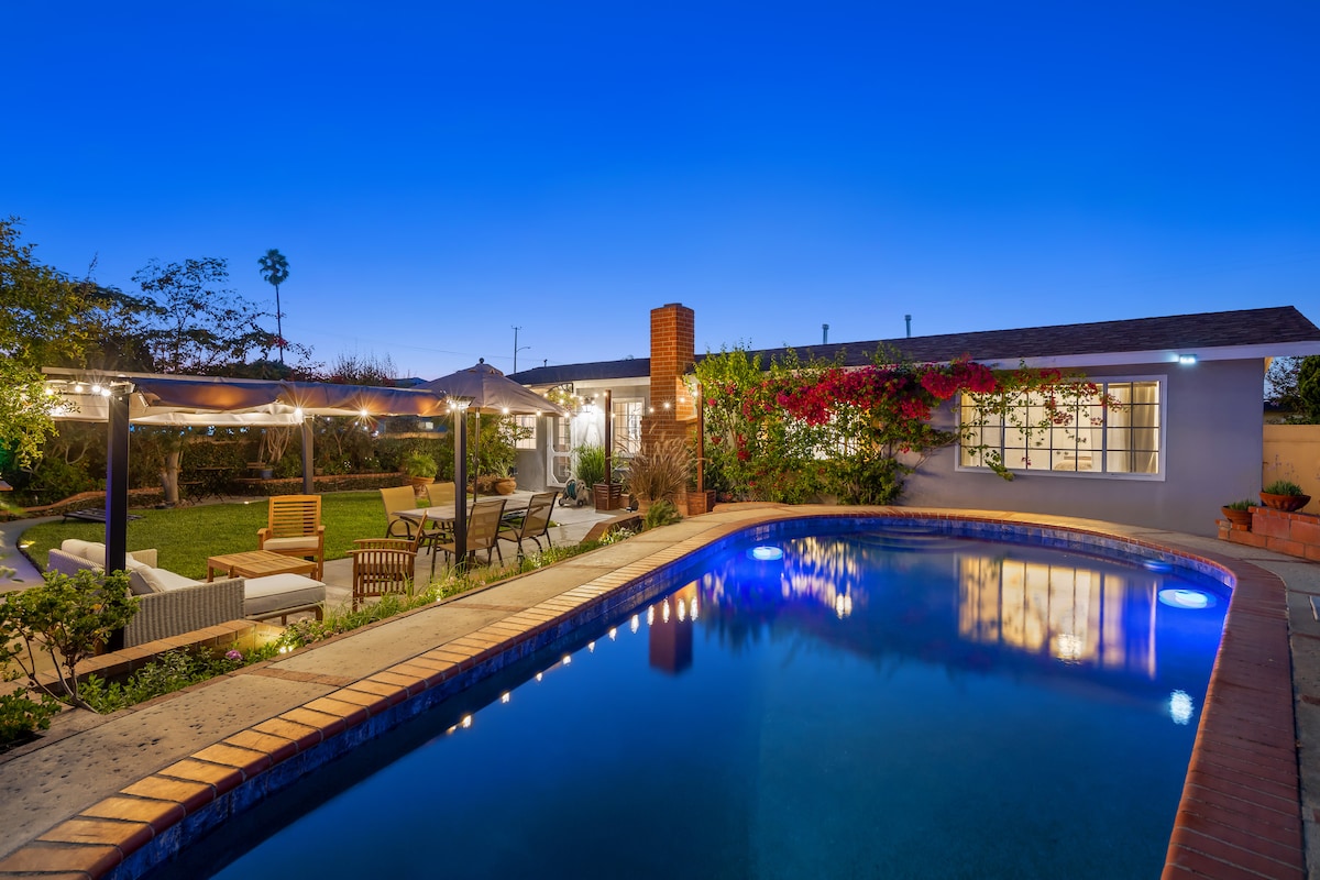 The backyard features a sparkling pool surrounded by a spacious patio area. Comfortable seating under a shaded cabana is seen, along with a dining set nearby. Lush greenery borders the space, and vibrant flowers are visible against the exterior of the home, adding a welcoming touch.