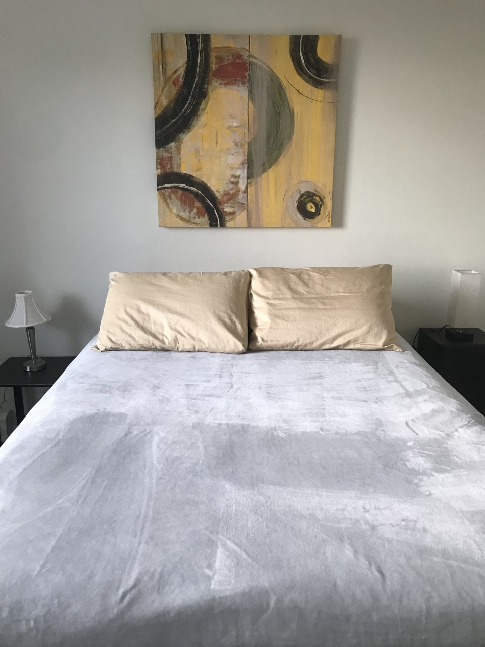 A spacious bed with soft beige linens is centered in the room, accompanied by two pillows. A light fixture stands on each side of the bed, while an abstract painting in yellow and dark tones hangs above, adding a pop of color to the neutral palette.