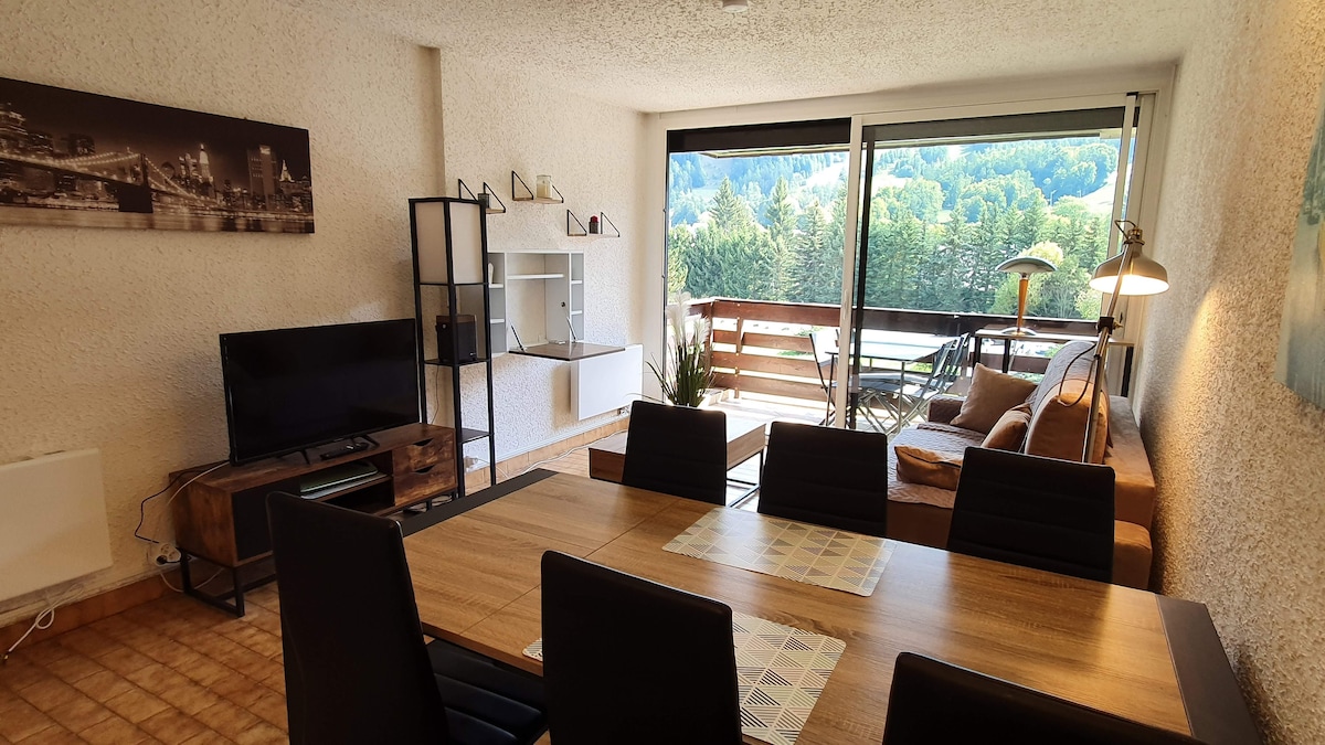 A contemporary living area features a dining table with dark chairs and a television unit. A large window provides a view of the surrounding greenery while the adjacent balcony includes outdoor seating. Soft lighting enhances the room's neutral tones.