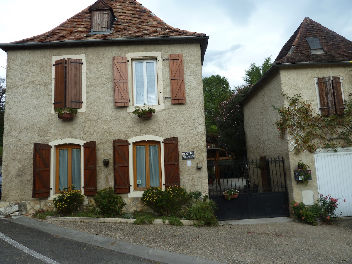 A charming two-story house with textured beige walls and a traditional slate roof is shown. Wooden shutters adorn the windows, with flower boxes adding color. A gated entrance leads to a small garden area that is visible in the front, showcasing well-maintained landscaping.