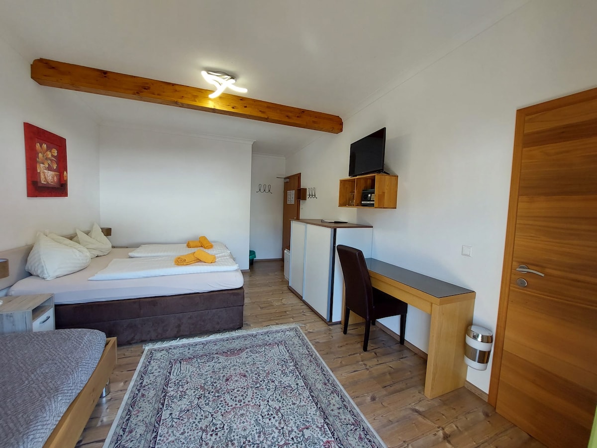 A spacious room features a split box-spring bed and a single bed, which can also function as a sofa. A small refrigerator, flat-screen TV, and desk are present. Wood beams accent the ceiling, and a warm wooden floor complements the decor.