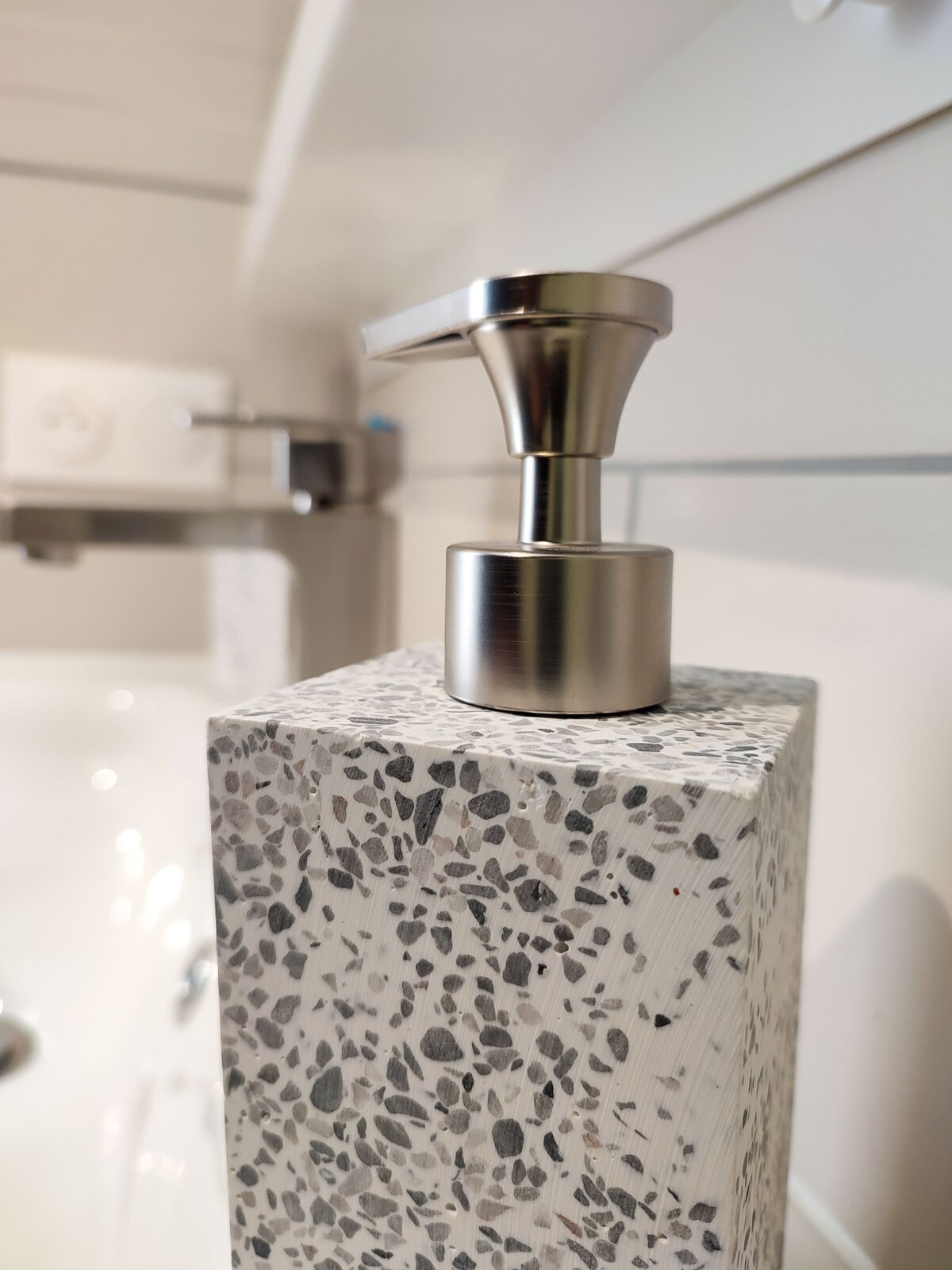A close-up of a modern soap dispenser is displayed, featuring a sleek metallic pump. The dispenser is mounted on a textured stone base with a speckled pattern, enhancing the contemporary design of the bathroom.