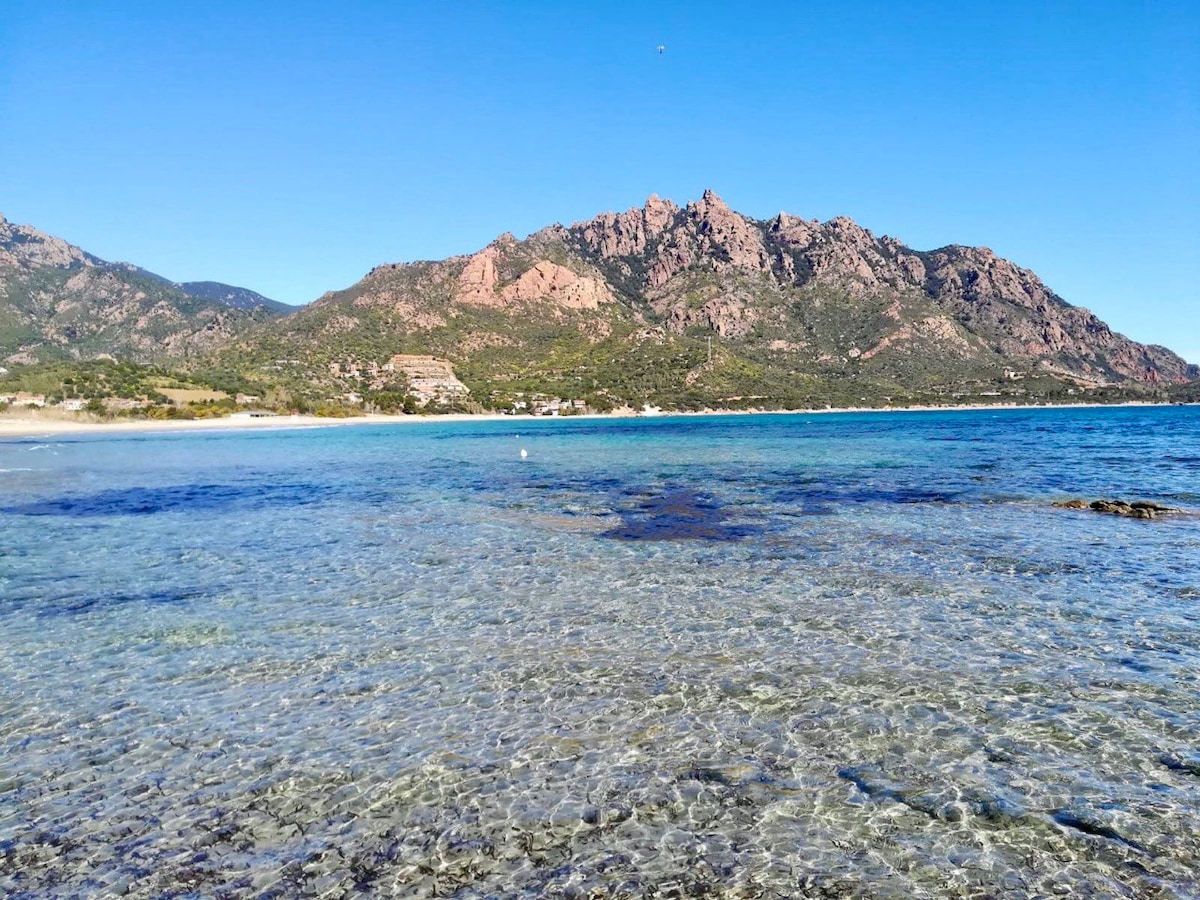 A clear view of the tranquil sea can be seen, with shallow water revealing smooth stones beneath. In the distance, a mountain range features rugged peaks and a mix of greenery along the shoreline, under a bright blue sky.