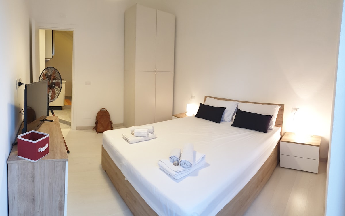 A bright and spacious bedroom features a large bed with white linens and dark accent pillows. Nearby, a wooden dresser holds a television, while two bedside tables accompany the bed. A standing fan is present for comfort, and closet space is available in the background.