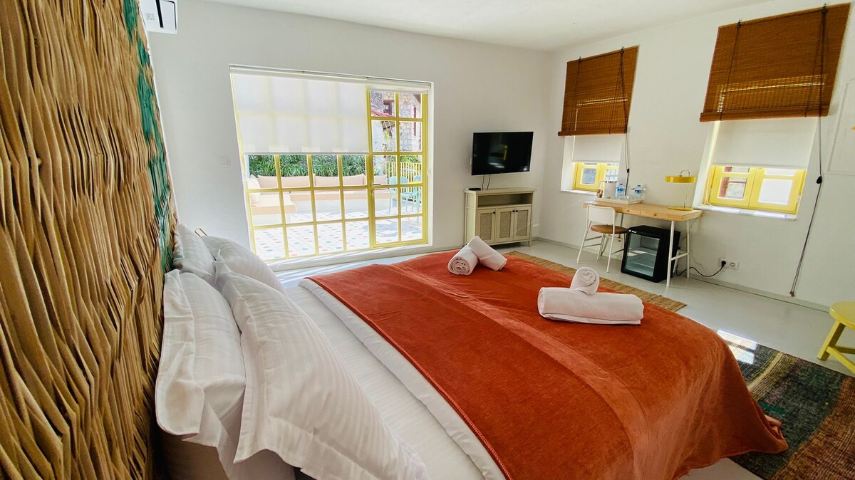 A bright and spacious sleeping area is presented, featuring a large bed with a rust-colored throw blanket and fluffy white towels. Natural light filters through window shades, and a flat-screen TV is mounted on the wall. Nearby, a small desk and chair provide a workspace.