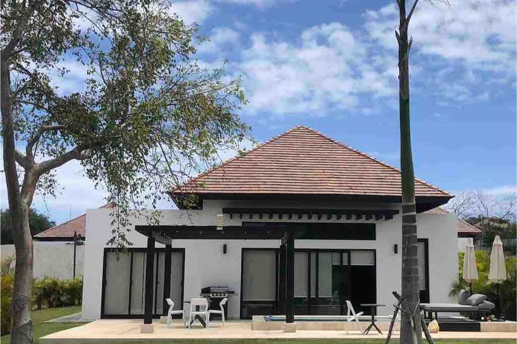 A modern villa is presented with a sloped tile roof and large glass doors that allow natural light to fill the interior. A spacious outdoor area features seating options and is surrounded by greenery, offering a peaceful atmosphere. The sky is clear with some scattered clouds.
