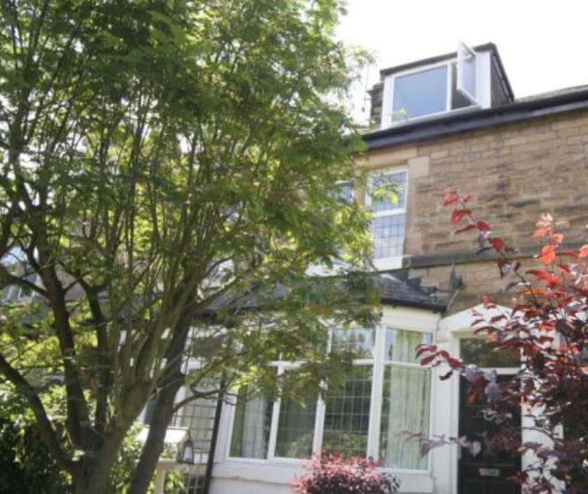 The property features a charming stone exterior framed by lush greenery. Large windows allow natural light to illuminate the entrance, while a quaint front garden with flowering plants adds a vibrant touch to the welcoming façade.