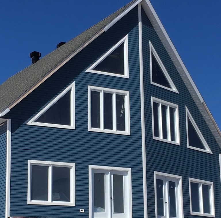 The exterior of a spacious blue duplex chalet is showcased, featuring triangular windows that enhance natural light. The structure is designed with a sloped roof and traditional siding, contributing to its distinctive appearance against a clear blue sky.
