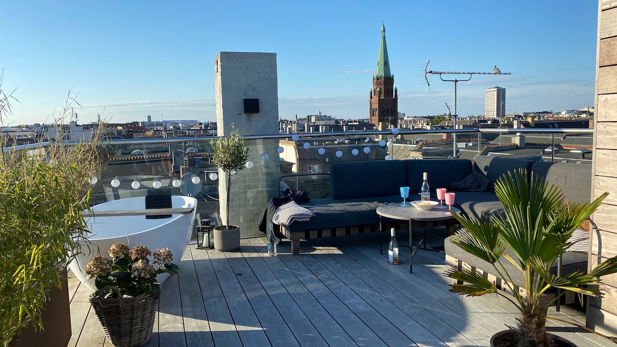 A spacious rooftop area features comfortable seating arrangements, a heated bathtub, and potted plants. The skyline of Copenhagen is visible in the background, showcasing the steeple of a nearby church and various city buildings under a clear blue sky.
