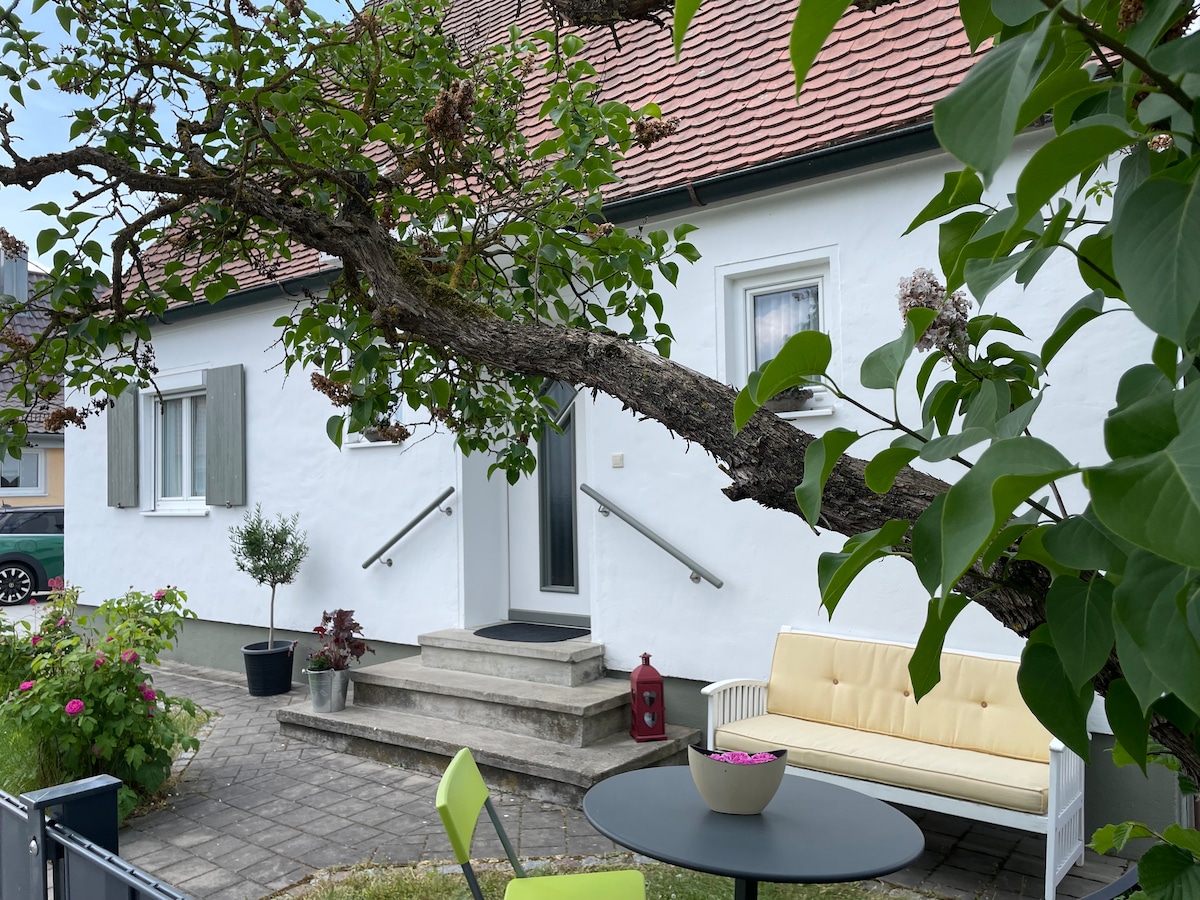 The exterior of the house is presented, featuring a small seating area with a light-colored sofa and a circular table. A flowering tree branches gracefully over the entrance, adding a touch of nature. Steps lead to the front door, framed by minimal landscaping.